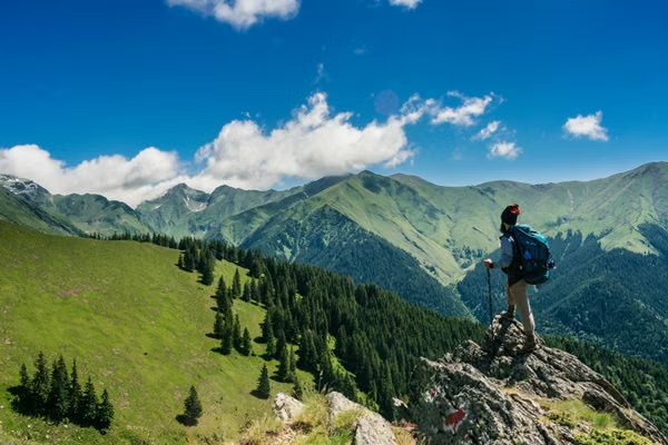A hiker with trekking poles and a backpack standing on a rocky summit, looking out over a wide mountain range